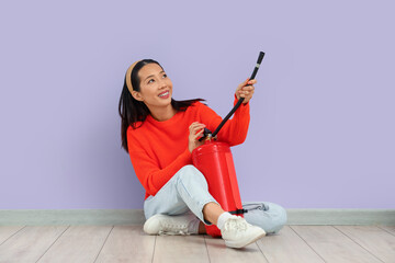Young Asian woman with fire extinguisher sitting against lilac wall
