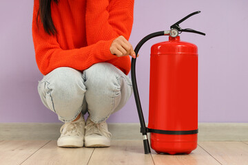 Young woman with fire extinguisher sitting against lilac wall