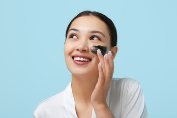 Young woman applying activated charcoal mask on her face against blue background
