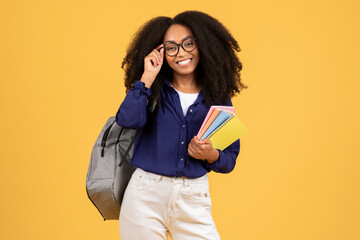 Happy black student with curly hair, backpack and copybooks, adjusting eyeglasses, poses on yellow...