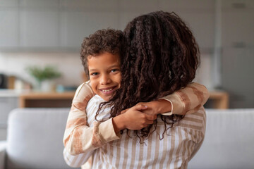 Smiling black boy with curly hair embracing his loving mother at home
