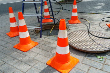 Orange cones installed around the dangerous area on the sidewalk. An open travel hatch. Laying of a new fiber-optic cable in the city network. orange construction cone - obrazy, fototapety, plakaty