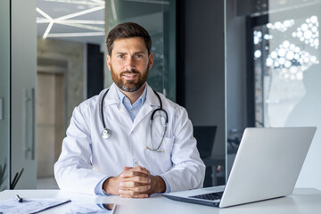 Portrait of a smiling confident male doctor in a white coat sitting at a desk in a clinic and looking at the camera