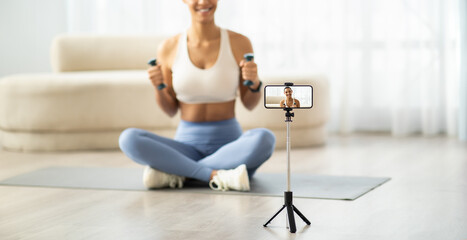 Smiling woman in sportswear holding dumbbells while recording a fitness video with a smartphone