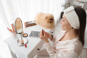 Young woman with cute Pomeranian dog doing makeup in bedroom