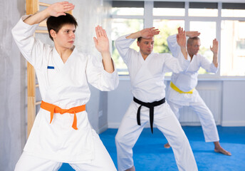 Concentrated young guy in white kimono practicing punches and blocks in gym during martial arts workout with male group. Shadow fight, combat sports training concept