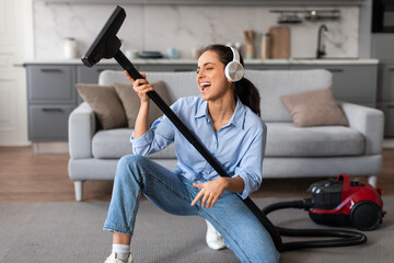 Joyful woman with headphones singing into vacuum cleaner