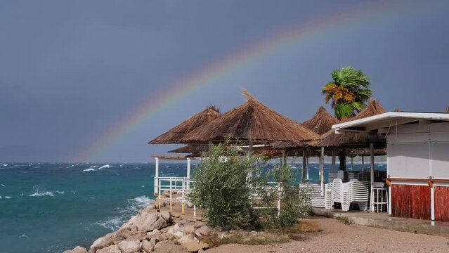 Slow motion colourful rainbow above the disquiet sea surface. Continuously changing weather conditions, light and shadow. Powerful waves crashing the coast after Storm Ciar&aacute;n smashed into Krk Island.