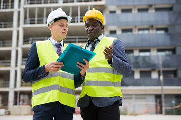 African-american and Eropean engineers standing on construction site and talking about new project. One man holding folder paper with documentation.