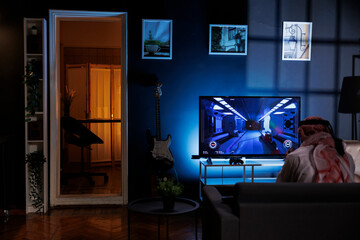 Rear-view shot of Muslim man seated on the sofa engrossed in playing video games. Image shows a...