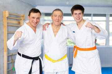 Three generations of men, older, middle-aged, and young, dressed in kimonos friendly embracing in training room. Concept of unity and traditions of family of martial arts fighters..