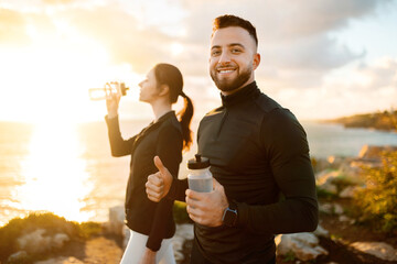 Active couple on coastal trail, man with water bottle giving thumbs up, woman drinking in background - obrazy, fototapety, plakaty