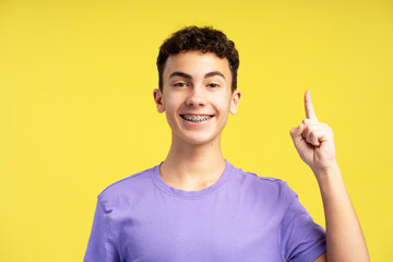 Smiling excited boy wearing casual purple t shirt looking at camera showing finger on copy space - obrazy, fototapety, plakaty