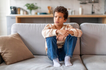 Pensive little boy with curly hair sitting on couch and looking away