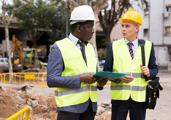 Two engineers, Caucasian and African-american men in hardhats and warnvests, standing and talking...
