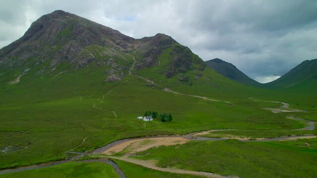 AERIAL: Stunning Scottish Highlands landscape with River Coe winding through green grassy valley. Remote charming white cottage on the river bank at the edge of moor under picturesque mountain peaks.