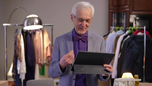A stylish elderly man in glasses, standing in a dressing room, holds a tablet with his hand and communicates on a video call. The man talks, adjusts his bow tie, gesticulates, nods and laughs.