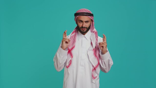 Arab person poses with fingers crossed and praying to allah over blank background, showcasing religion and spiritual devotion. Man in muslim clothes being religious, worshipping God with faith.