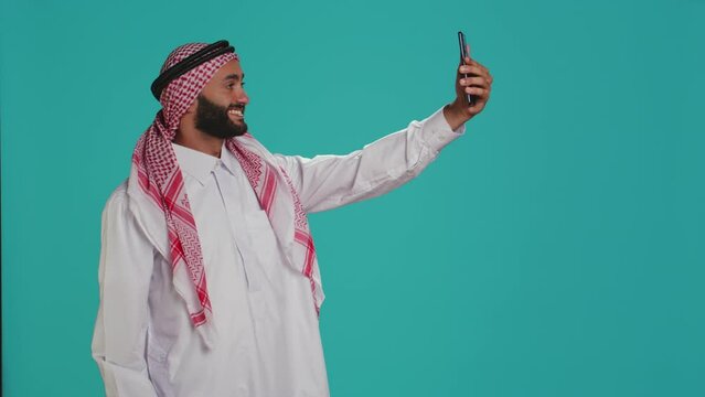 Arab man takes pictures with smartphone, laughing in studio using smartphone in front of studio camera. Middle eastern person with traditional headscarf taking photos in cultural attire.