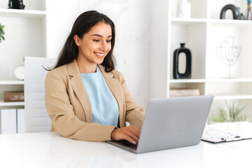 Happy young adult indian or arabian business woman entrepreneur, wearing suit, sitting in the office, using laptop at work, working on a project at workplace, searching information in internet, smiles