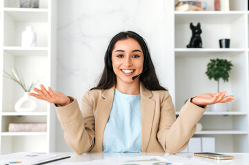 Successful attractive arabian or indian woman with headset, female teacher, business consultant sitting in the office during online consultation with client, recording video tutorial, smiles at camera