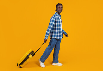 Smiling young black man casually walking with yellow suitcase on orange background