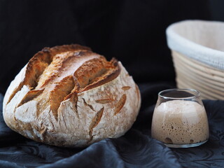 Fresh sourdough starter in a glass jar with loaf and banneton on black fabric