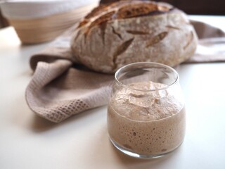 Closeup on fresh sourdough in a glass jar, bread and banneton in background, white table