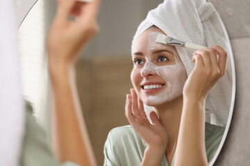 Woman applying face mask in front of mirror indoors. Spa treatments