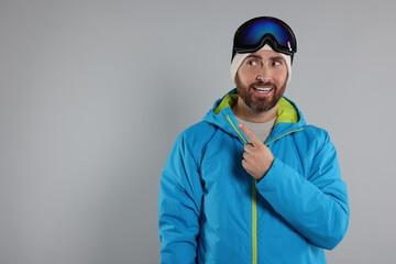 Winter sports. Happy man in ski suit and goggles pointing at something on gray background, space for text