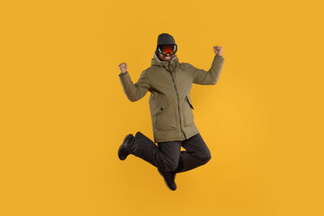 Winter sports. Happy man in ski suit and goggles jumping on orange background
