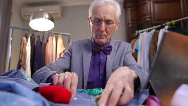 A man marks a line with chalk, using a measuring tape, on the fabric of a blue shirt. An elderly gray-haired fashion designer wearing glasses works at a table in the workshop of his home.