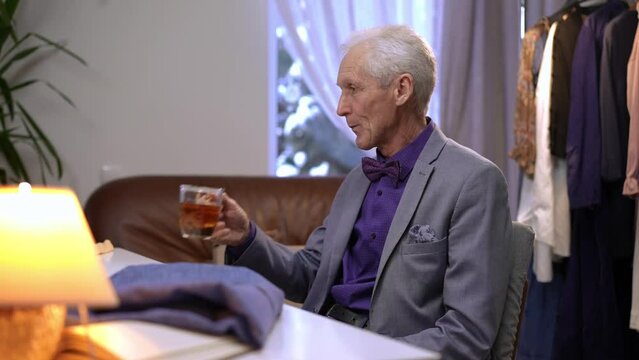An elderly male fashion designer sits in his office at the table and drinks tea from a glass cup