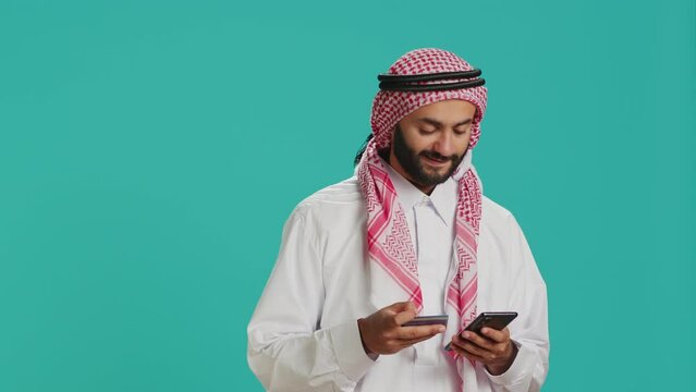 Smiling adult using card to pay online while shopping on internet store website with smartphone. Middle eastern guy in islamic clothing making baking transfer to buy something on sale.