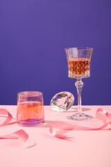 Front view of a wine glass and a cup decorated on pink surface. A diamond and some pink ribbons featured. Alcohol drink, cocktail party concept