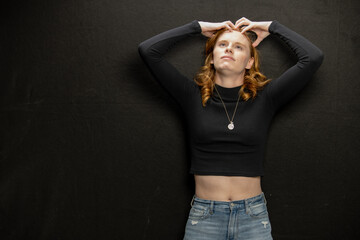 Young woman with red hair in black sweater and jeans poses in studio before a black background