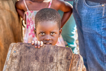 portrait of an african girl in the village , hiding behind a wooden board, people in the background