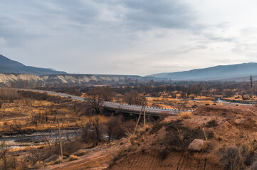 Fototapeta premium Panorama of a mountain valley, road and bridge over a river in a mountainous countryside. Brown earth and bald trees.