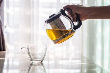 hands pouring tea from transparent kettle to teacup on windows background with copy space.