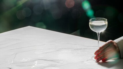 A person's hand resting on a marble surface next to a wine glass filled with white wine, in a dimly...