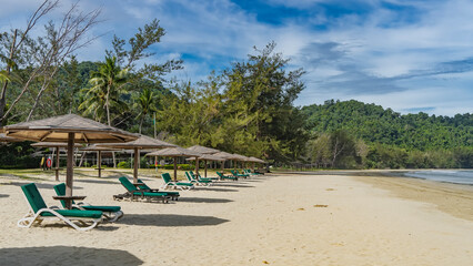 Sun loungers stand in a row on the sandy beach by the ocean in the shade of sun umbrellas. A green hill against a background of blue sky and clouds. Malaysia. Borneo. Kota Kinabalu