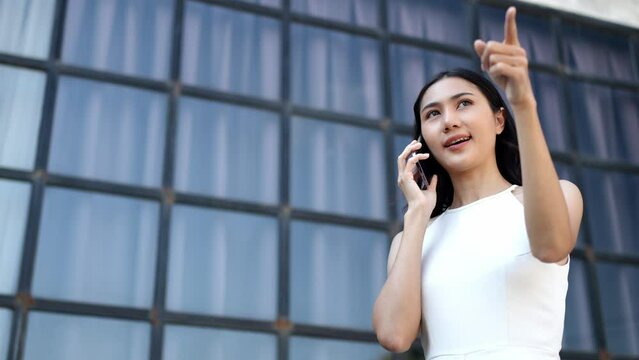 A confident Asian businesswoman stands talking on the phone with her business partner in the office outside outdoors using an application on a mobile phone fast connection online communication concept