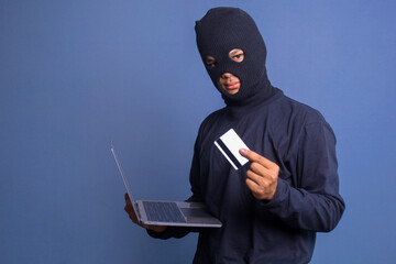 Computer hacker with balaclava holding credit card and stealing data from a laptop. Concpet of...