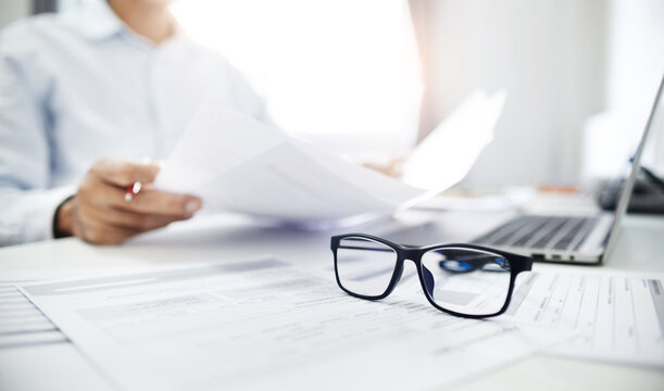 Close-up shot at eyeglasses, blurred businessman review many documents placed on the desk and using laptop meeting online with business group to plan and start new business