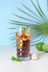 Glass of cold Cuba Libre cocktail, palm leaf and seashell on white table