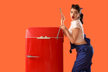 Shocked young pin-up worker with wrenches and retro fridge on orange background