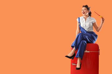 Attractive pin-up worker with screwdriver sitting on retro fridge against orange background