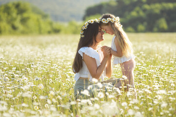 Portrait of tender mother and daughter happy together, hugging in nature in a field of blooming chamomile flowers on a summer day. Mom and girl with long hair and daisy wreath, cotton casual wear 