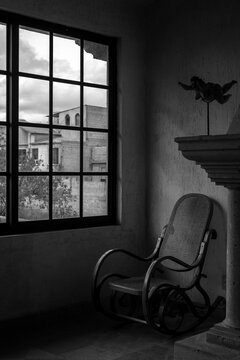 Vieja silla mecedora de madera y paja tostada trenzada, al lado de una ventana. Blanco y negro.