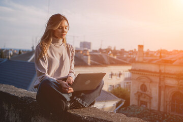 Woman freelancer uses laptop on cement wall outdoors against the sky and the roof of the city. The...